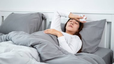 Asyalı bir kadın kendini hasta hissediyor ve yatak odasında öksürürken ve hapşırırken peçeteyi elinde tutarken ve alnına dokunurken yatakta uyuyor.