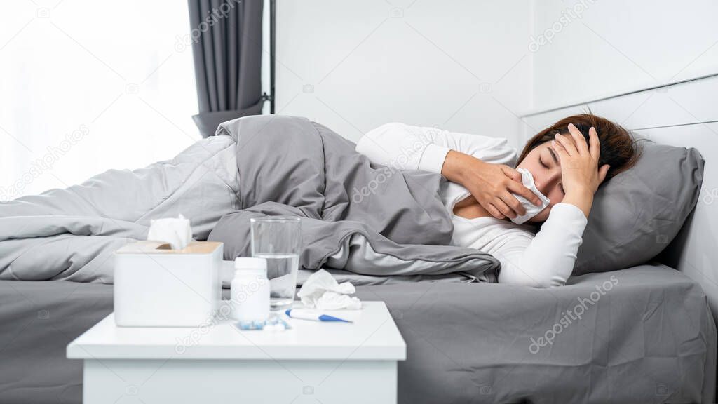 Asian woman using tissue to cough, sneezing and touching forehead while feeling sick and lying in blanket on the bed after using thermometer to checking body temperature and take medicine in bedroom