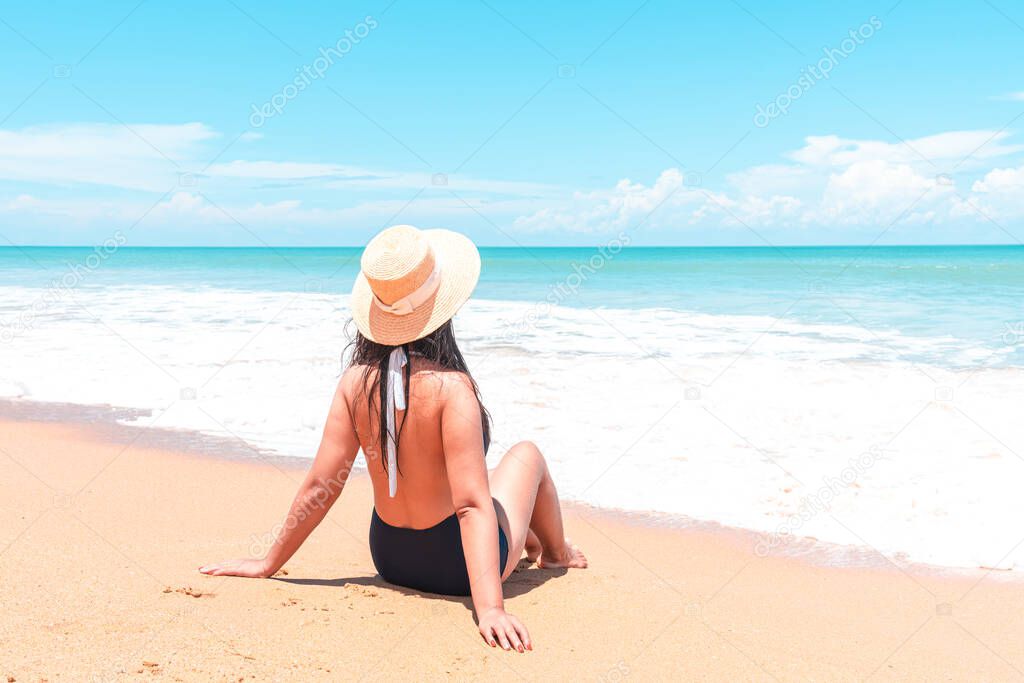Back view of asian traveler woman in swimsuit and straw hat to sitting in the spray of the sea to looking view the blue horizon on the sea while relaxing in holiday trip