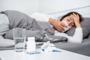 Vücut ısısını ölçmek ve ilaç almak için termometre kullandıktan sonra hasta hissedip yatağa uzanan Asyalı kadın, öksürmek, hapşırmak ve alnına dokunmak için ağzını kapatır.
