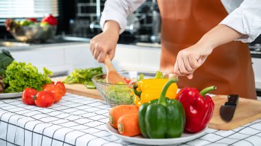 Asyalı ev hanımı önlük takıyor ve kepçeyle kasede sebze salatasını karıştırırken kahvaltıda, öğle yemeğinde ve akşam yemeğinde evde sağlıklı yemek hazırlıyor.