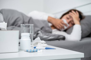 Asyalı kadın öksürürken, hapşırırken ve alnına dokunurken termometre kullanıp vücut ısısını ölçtükten sonra kendini hasta hissederken ve yatak odasında ilaç alırken mendil kullanıyor.
