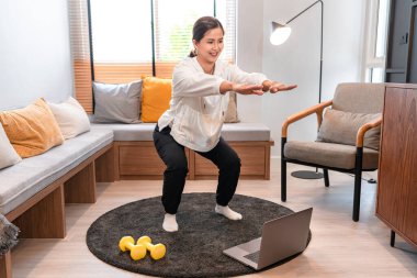 Antrenmandan önce kollarını ve bacaklarını açan Asyalı bir kadın ve evinde yoga dersi vermek için dizüstü bilgisayar kullanıyor.