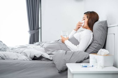 Asyalı kadın öksürmek, hapşırmak ve ilaç almak için bir bardak su tutarken kendini hasta hissedip vücut ısısını ölçmek için termometreyi kullandıktan sonra battaniyeye oturmuş.