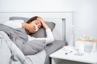 Asyalı kadın öksürürken, hapşırırken ve alnına dokunurken termometre kullanıp vücut ısısını ölçtükten sonra kendini hasta hissederken ve yatak odasında ilaç alırken mendil kullanıyor.