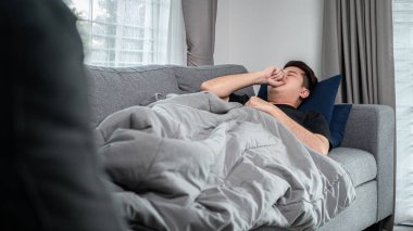 Asyalı bir adam, yüksek ateş, baş ağrısı ve öksürük ve hapşırma sırasında elini kullanarak evdeki kanepeye uzanıp dinlenir.