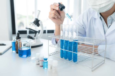 Tıbbi yüz maskesi takan bilim kadını test tüpüne solüsyon atmak ve laboratuvarda mikroskopla çalışırken deneysel koronavirüs aşısı için reaksiyon aramak için damlatıcı kullanıyor.