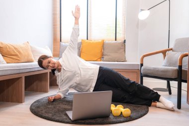 Asyalı kadın yoga egzersizi yapıyor, kolunu kaldırıyor ve dizüstü bilgisayarla evdeki oturma odasında yoga dersi veriyor.