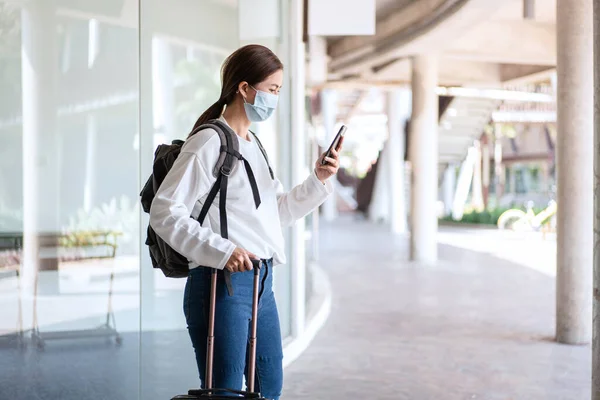 Tourist woman with luggage using smartphone to searching destination while wearing medical face mask to protection from coronavirus and travel with new normal lifestyle during covid-19 outbreak