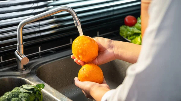 Hands of asian housewife washing orange and vegetables with water in sink while wearing apron and preparing to cooking for healthy meal in the kitchen at home