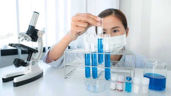 Scientist woman in medical face mask is holding test tube with blue liquid and looking reaction of experimental coronavirus vaccine while working with microscope in laboratory