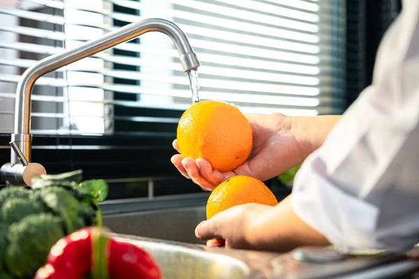 Hands of asian housewife washing orange and vegetables with water in sink while wearing apron and preparing to cooking for healthy meal in the kitchen at home