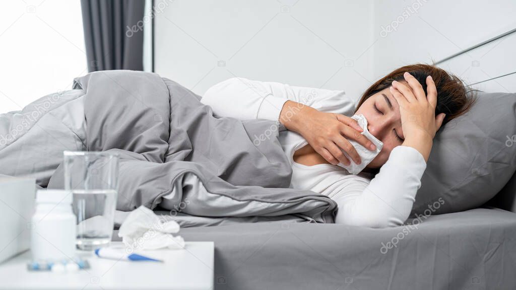 Asian woman using tissue cover your mouth to cough, sneeze and touching forehead while feeling sick and lying in blanket on the bed after using thermometer to check body temperature and take medicine