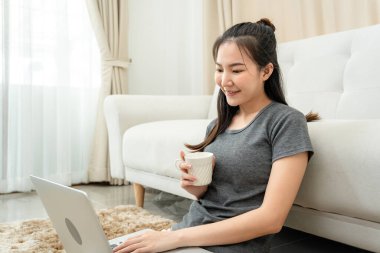 Asyalı serbest çalışan bir kadın kahve fincanı içerken ve dizüstü bilgisayarını kullanarak müşteriden sipariş almak için yerde otururken ürünleri kontrol etmek için merkez ofisteki çevrimiçi pazarlama sırasında...