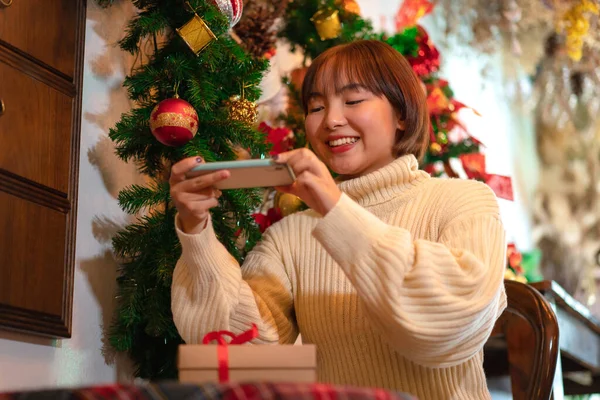 セーターを着た美しい女性は笑顔で クリスマスツリーの前でギフトボックスの写真を撮るためにスマートフォンを使用しています — ストック写真