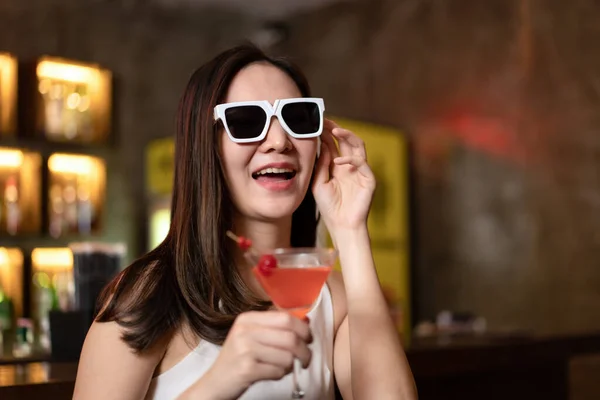 Beautiful woman is wearing sunglasses and drinking cocktail during having enjoy to relaxing while sitting alone to listening music on counter bar in restaurant or hotel bar