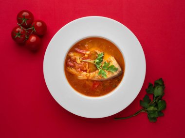 Beyaz tabakta balık çorbası, taze domates ve kırmızı arka planda maydanoz. Kopyalama alanı ile genel görünüm.