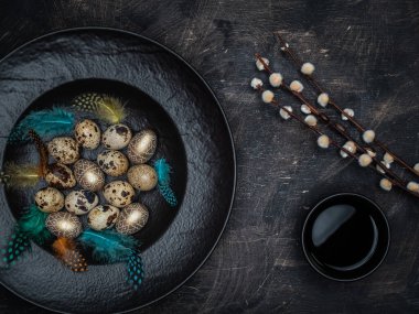 Easter concept. Quail eggs, golden colored eggs and colorful feathers in black bowl. Cup of black tea and pussy willow on vintage background.