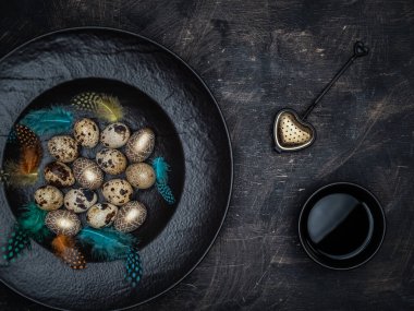 Paskalya konsepti. Bıldırcın yumurtaları, altın renkli yumurtalar ve siyah kasedeki renkli tüyler. Bir fincan siyah çay ve çay süzgeci. Kopyalama alanı ile genel görünüm.
