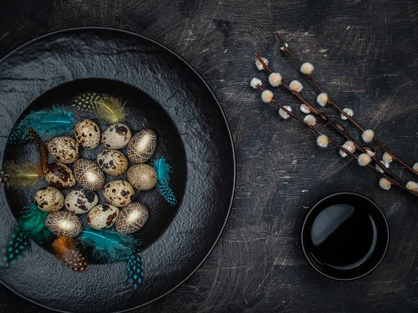 Easter concept. Quail eggs, golden colored eggs and colorful feathers in black bowl. Cup of black tea and pussy willow on vintage background.