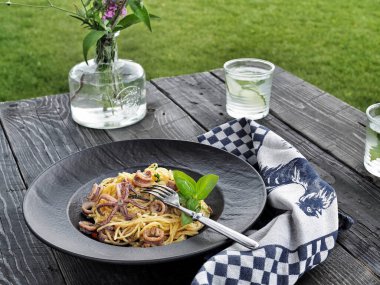 Bahçedeki ahşap masada siyah tabakta deniz ürünleri makarnası. Kalamarlı spagetti. Arka planda limonlu su. Yatay çekim.