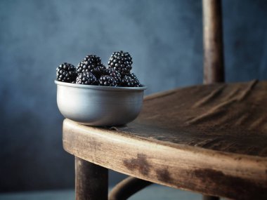 Eski ahşap sandalyede metal kasede taze böğürtlen. Uzayı kopyala Karanlık ve karamsar. 
