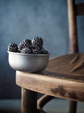 Eski ahşap sandalyede metal kasede taze böğürtlen. Uzayı kopyala Karanlık ve karamsar. Dikey çekim.