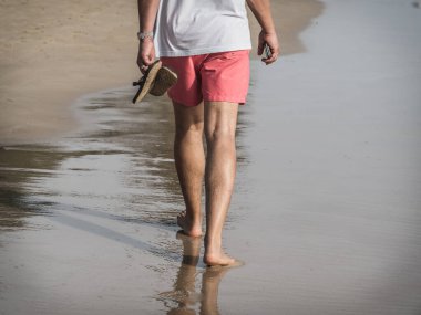Kumsalda yürüyen, kırmızı şortlu, akıllı telefonlu beyaz bir adam. Yalınayak ayaklarının yansıması..