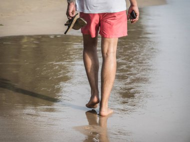 Kumsalda yürüyen, kırmızı şortlu, akıllı telefonlu beyaz bir adam. Yalınayak ayaklarının yansıması..