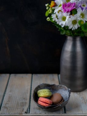 Metal vazoda bir buket çiçek ve ahşap masadaki kasede üç renkli kurabiye. Uzayı kopyala Karanlık ve karamsar. 