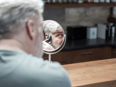 Kıdemli beyaz adam masa aynasına bakar ve beyaz saçlarını elektrikli makasla keser. Evde kendi kendini buduyor. Yatay çekim. Boşluğu kopyala.