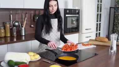 Önlüklü genç bir kadın mutfakta domatesli omlet, tavaya yumurtalı vişneli domates ekliyor.