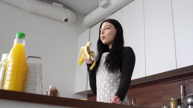 Önlüklü genç bir kadın mutfakta muz yer, kız evde muz yer ve meyve yer.