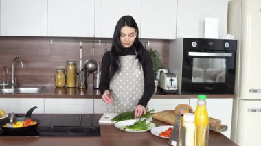 Önlüklü genç bir kadın mutfakta yeşil soğan, dereotu ve maydanoz doğruyor.