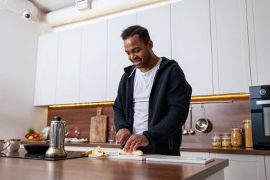 Beyaz mutfakta muz kesen, evde yemek yapan ve gülümseyen Afro-Amerikan adam.