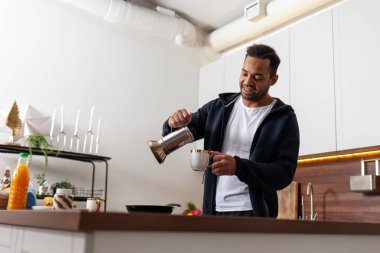 Afro-Amerikalı adam mutfakta kahve yapıyor ve gülümsüyor, adam evde kahvesini fincana dolduruyor.