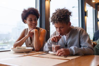 Afro-Amerikalı kadın ve oğlu kafede oturup kahve içiyorlar ve gülümsüyorlar, kıvırcık saçlı genç ve annesi restoranda öğle yemeği yiyorlar.