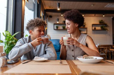Afro-Amerikalı kadın ve oğlu kafede oturup kahve içiyorlar ve gülümsüyorlar, kıvırcık saçlı genç ve annesi restoranda öğle yemeği yiyorlar.
