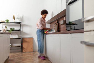 Afro-Amerikalı kadın mutfak lavabosunda bulaşık yıkıyor ve gülümsüyor, kız evi temizliyor ve beyaz mutfağı temizliyor.