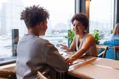 Afro-Amerikalı bir kadın ve oğlu kafede oturmuş kıvırcık saçlı bir ergenle konuşuyorlar ve annesi restoranda oturmuş gülümsüyor.