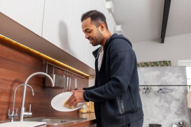 Afro-Amerikalı adam mutfakta tek başına bulaşık yıkıyor, evi temizliyor ve ev işi yapıyor.