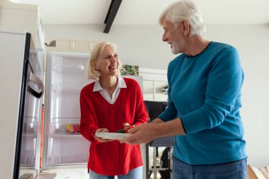 Yaşlı çift mutfakta buzdolabı kullanıyor, gri saçlı kadın ve kocası mutfakta yemek pişiriyor ve buzdolabından sebze alıyorlar.
