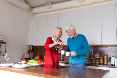 Yaşlı bir çift mutfakta kahve içip gülümsüyor, yaşlı kadın ve erkek ellerinde çay fincanları tutuyor ve evde sarılıyorlar.