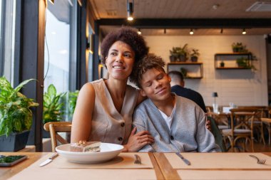 Afro-Amerikalı bir kadın ve oğlu kafede oturmuş kıvırcık saçlı bir ergen ve annesi restoranda oturmuş birbirlerine sarılıyorlar.