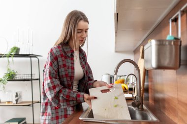 Mutsuz genç bir kadın mutfak lavabosunda bulaşık yıkıyor, üzgün bir kız evi temizliyor ve beyaz mutfağı toparlıyor.