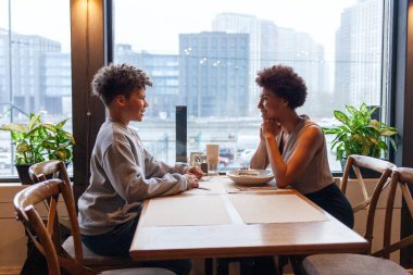 Afro-Amerikalı bir kadın ve oğlu kafede oturmuş kıvırcık saçlı bir ergenle konuşuyorlar ve annesi restoranda oturmuş gülümsüyor.