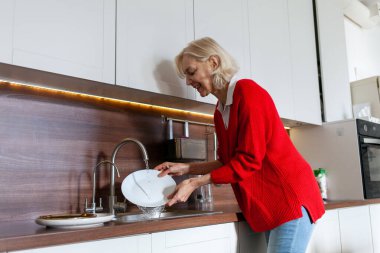 Kırmızı bluzlu yaşlı kadın mutfakta bulaşıkları yıkıyor, gri saçlı büyükanne ev işi yapıyor ve beyaz mutfakta tek başına temizlik yapıyor.
