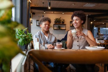 Afro-Amerikalı kadın ve oğlu kafede oturup kahve içiyorlar ve gülümsüyorlar, kıvırcık saçlı genç ve annesi restoranda öğle yemeği yiyorlar.