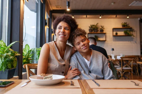 Afro-Amerikalı bir kadın ve oğlu kafede oturmuş kıvırcık saçlı bir ergen ve annesi restoranda oturmuş birbirlerine sarılıyorlar.