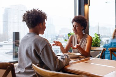 Afro-Amerikalı bir kadın ve oğlu kafede oturmuş kıvırcık saçlı bir ergenle konuşuyorlar ve annesi restoranda oturmuş gülümsüyor.